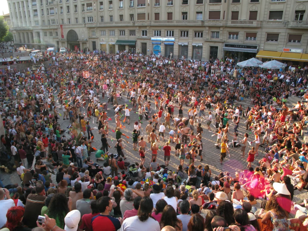 Many jugglers standing on a town square competing in a game while a huge audience surrounds them and watches